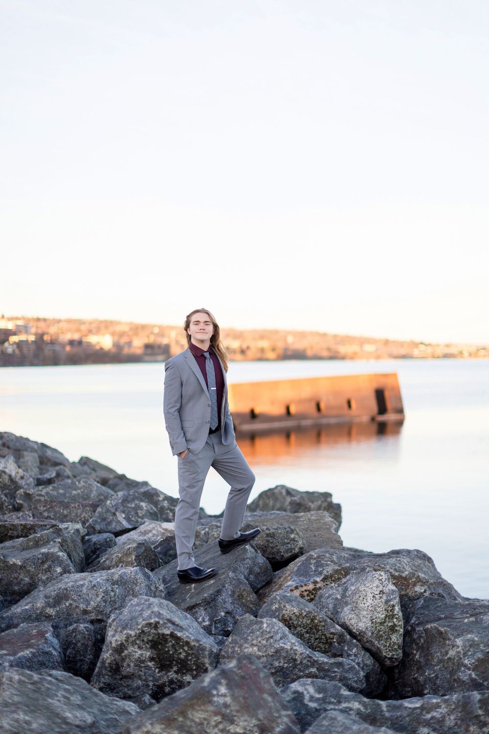 Person in gray suit walking across rocky shoreline during sunset with water and horizon in background.
