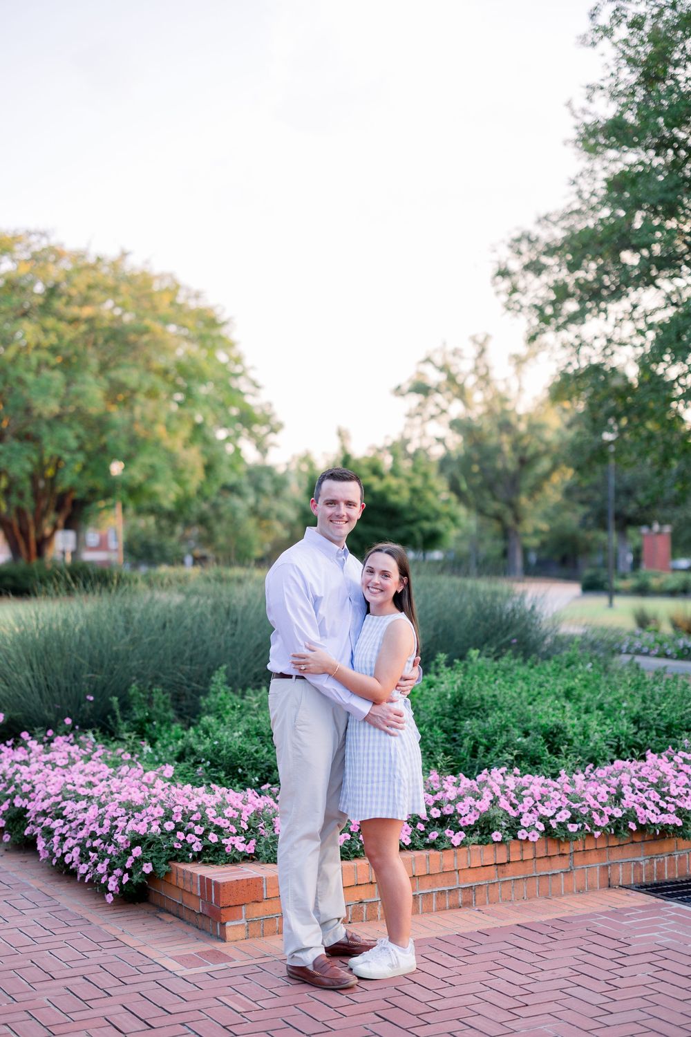 A couple poses together in a garden with pink flowers and brick landscaping on a sunny day.