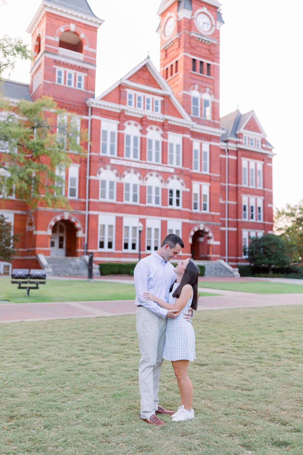 A couple shares a romantic moment on the lawn in front of a historic red brick university building with Victorian architecture.