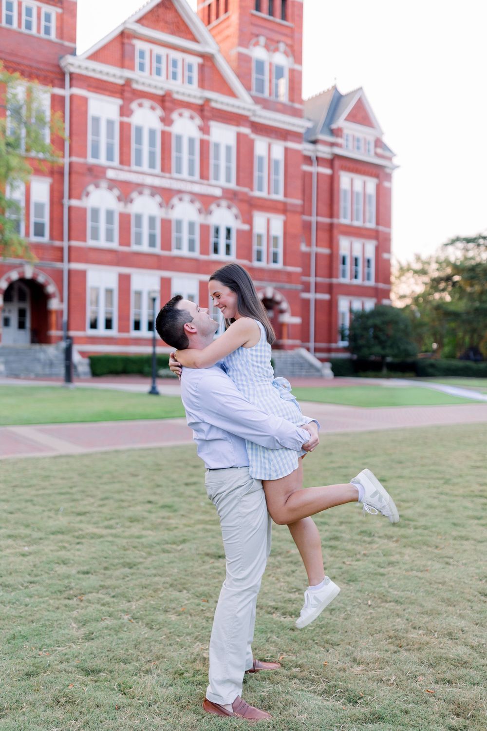 A romantic couple embraces on a lawn in front of a grand red brick Victorian-style building during sunset.