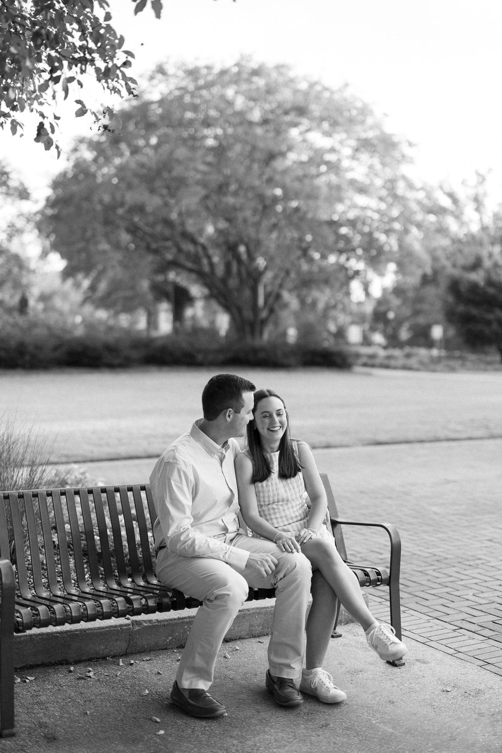 A couple playfully interacts while sitting on a park bench in a black and white photograph.