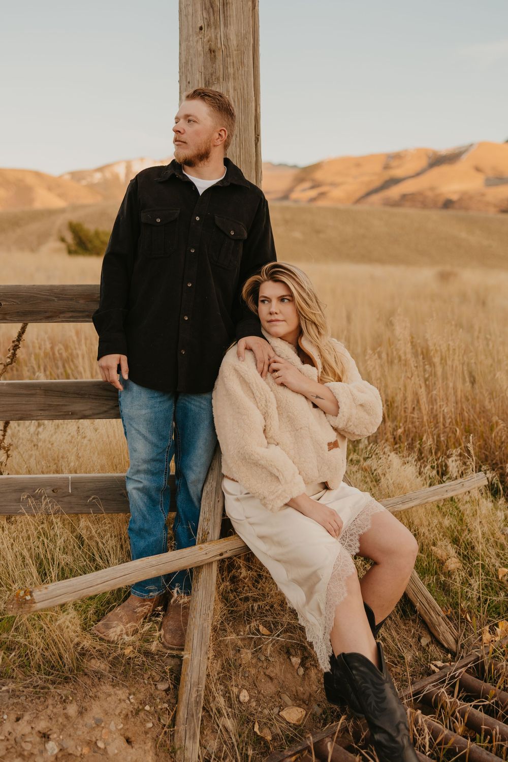 A couple sits together on a wooden fence at sunset in a golden field with mountains in the background of Paradise Valley, Montana