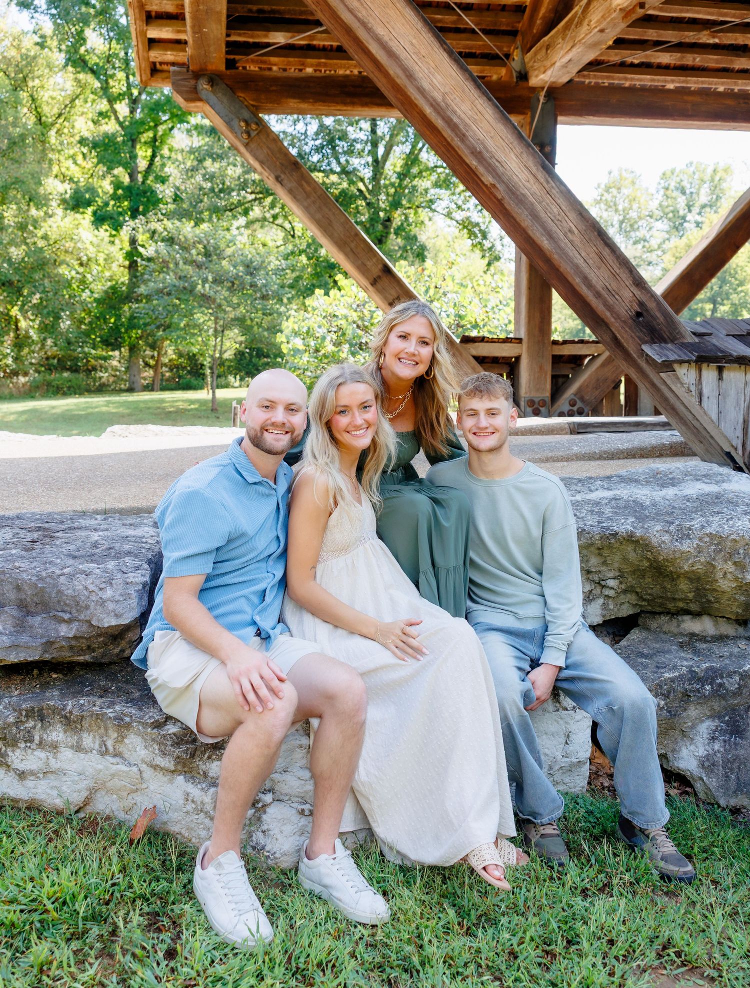Group sitting together on rustic wooden covered bridge in casual summer outfits enjoying outdoor photo session.