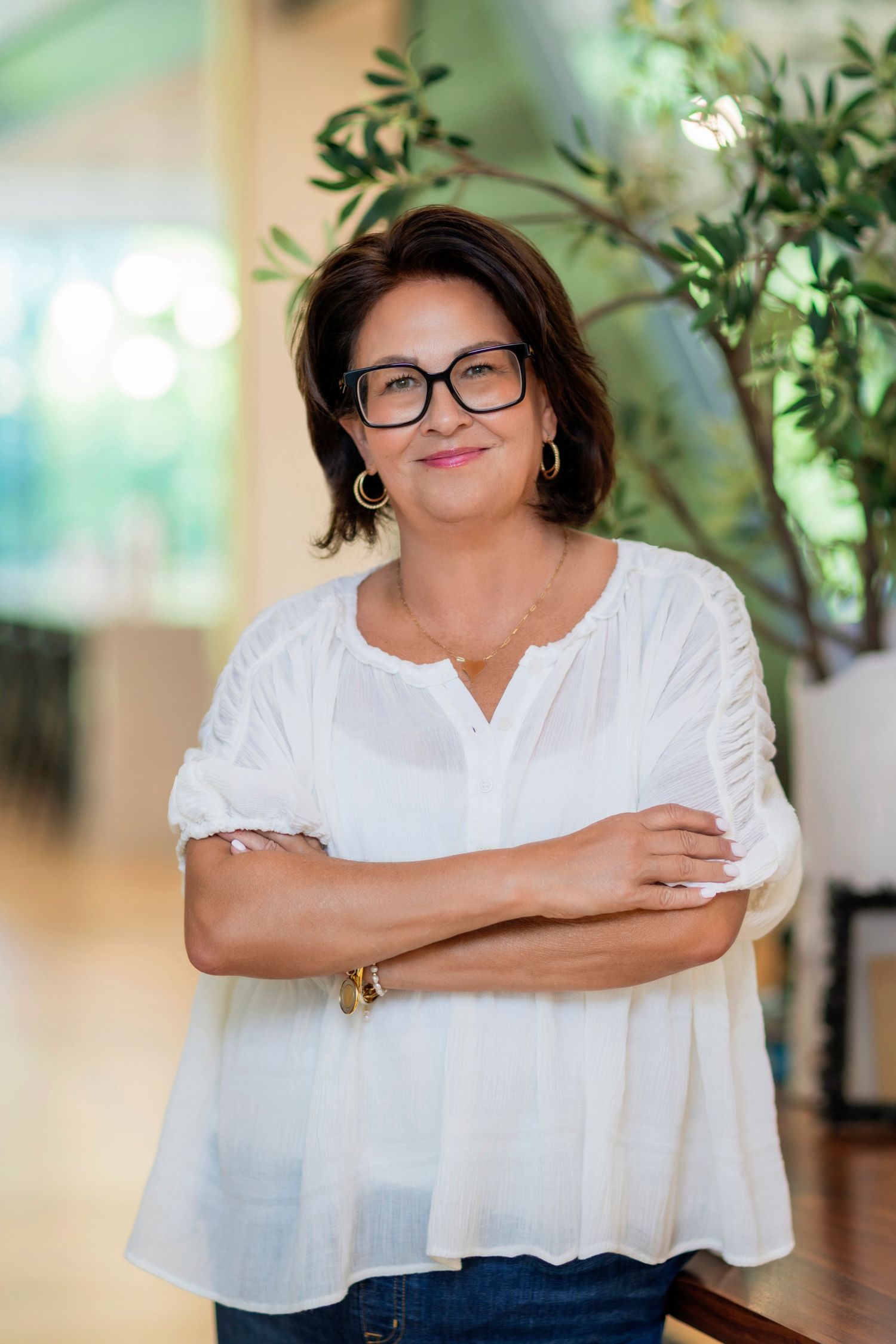 A person in a white button-down shirt smiles warmly while posing in front of a blurred green plant background.