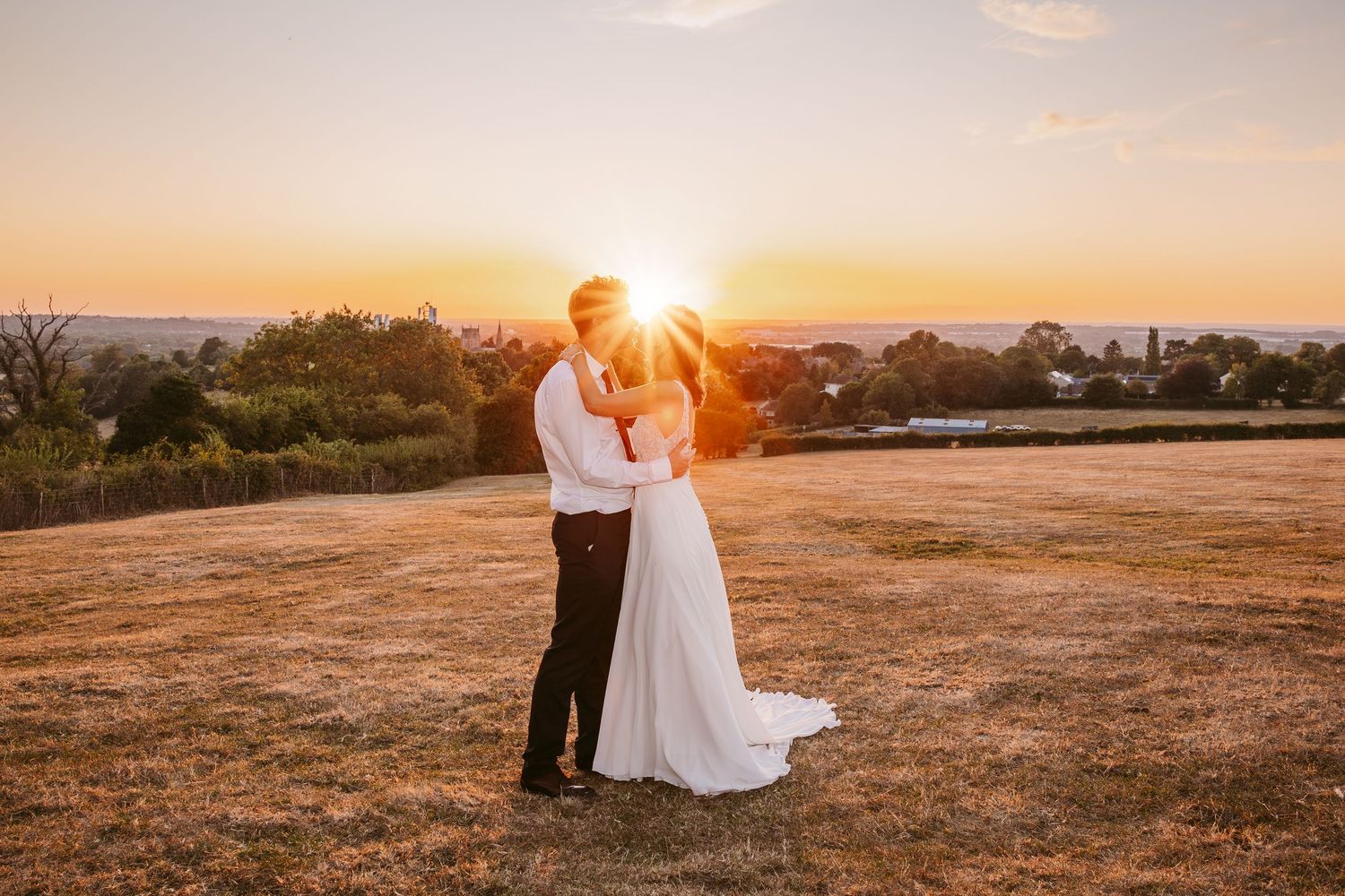 Newlyweds share intimate moments against a dramatic sunset backdrop in an open field.