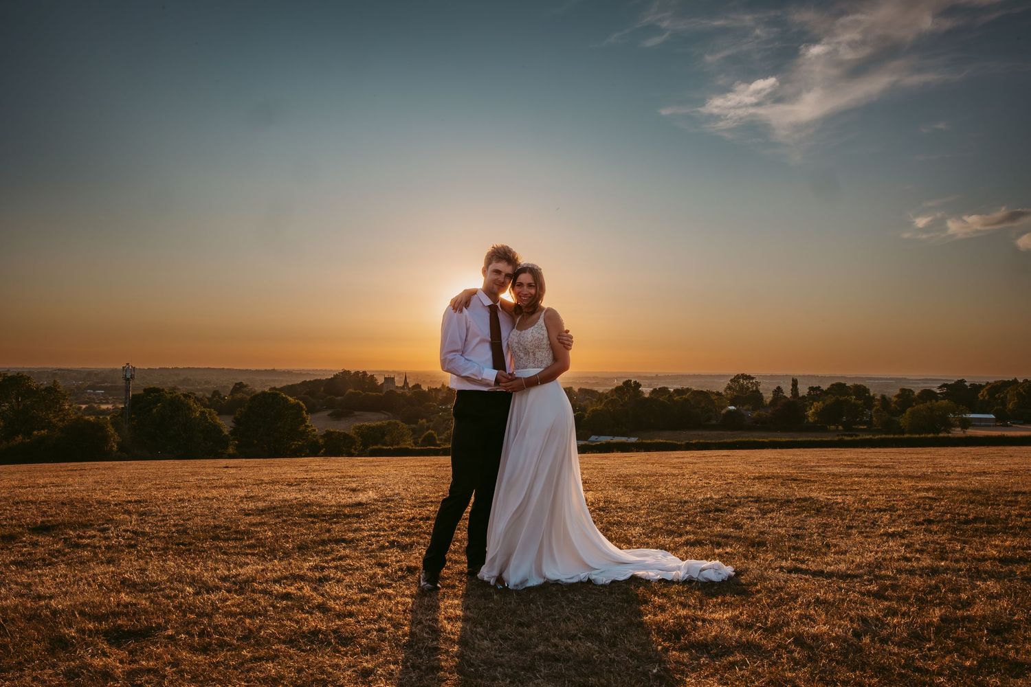 A wedding couple embraces in silhouette against a dramatic sunset sky in a rural field.