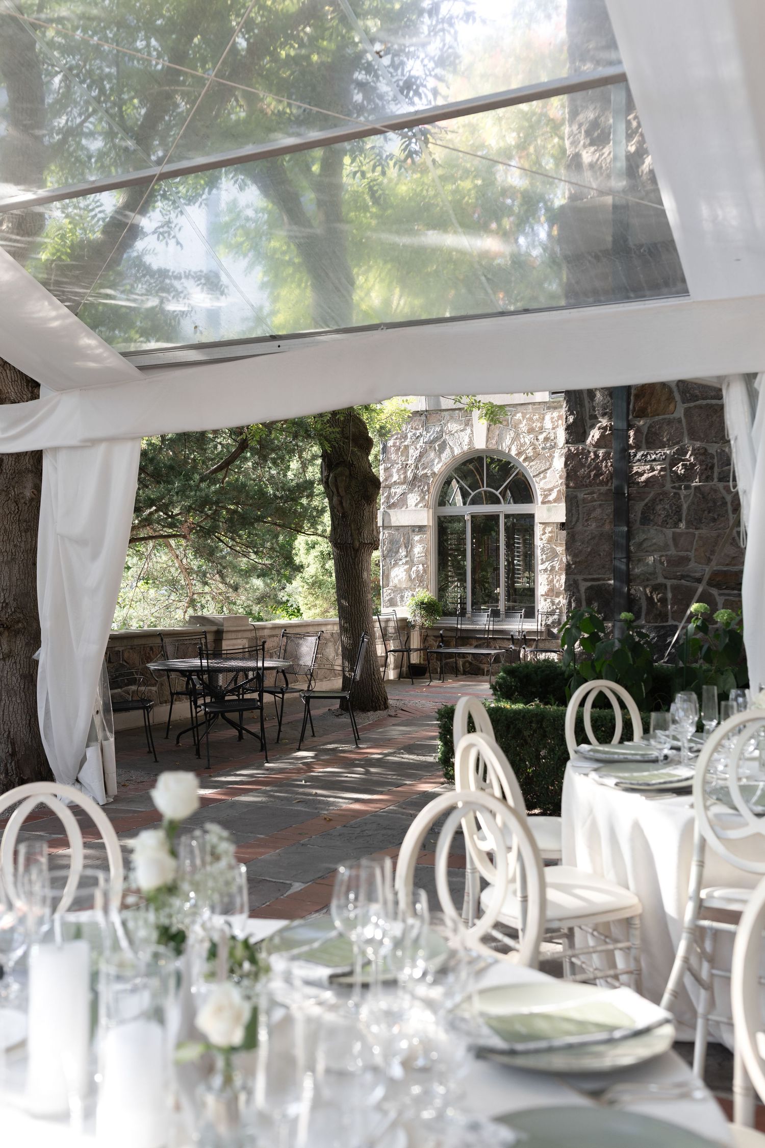 Elegant outdoor wedding reception setup with white tablecloths, place settings and flowing sheer curtains in a covered patio area.