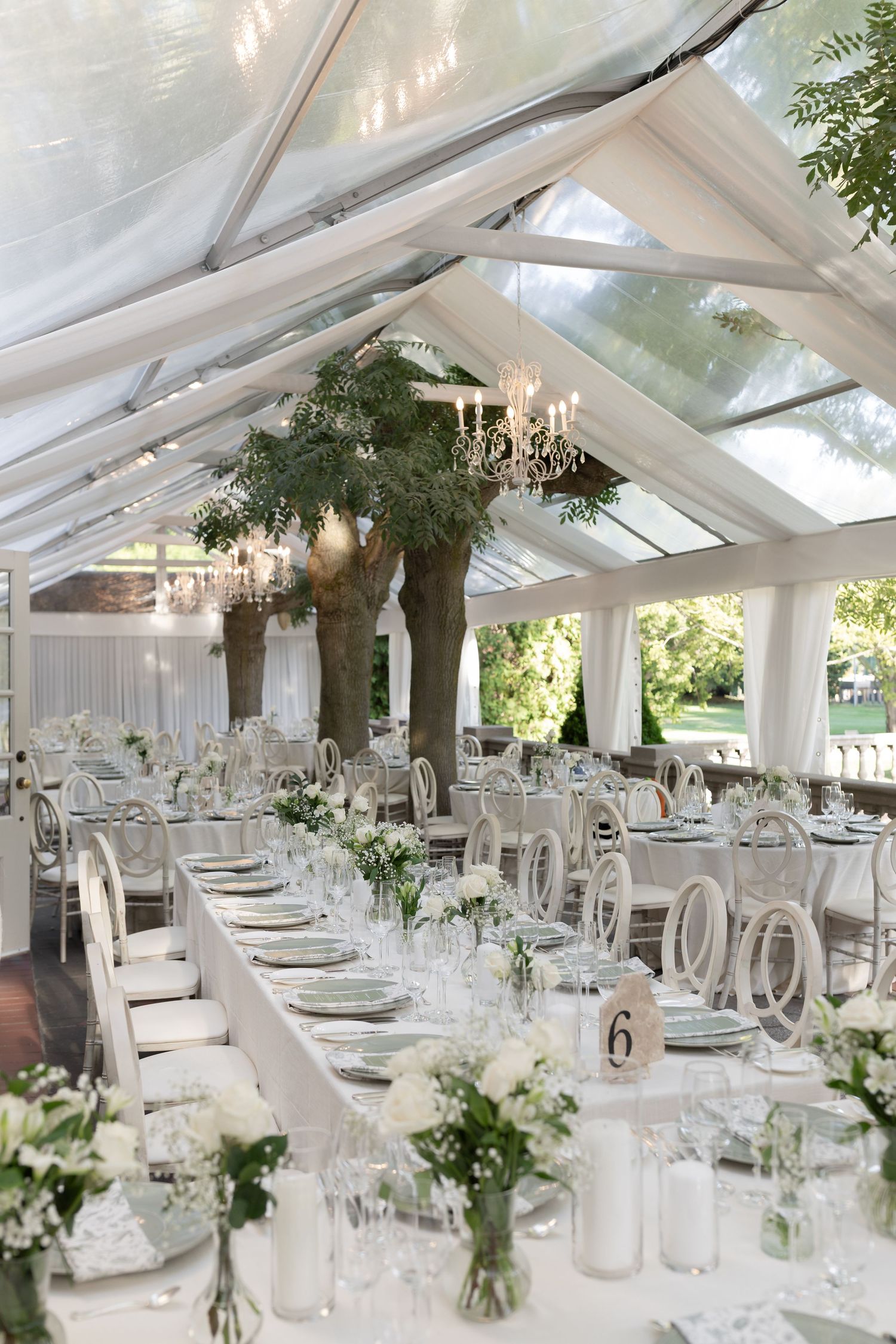 Elegant white wedding reception tent with crystal chandeliers, draped fabric, and greenery centerpieces on long dining tables.