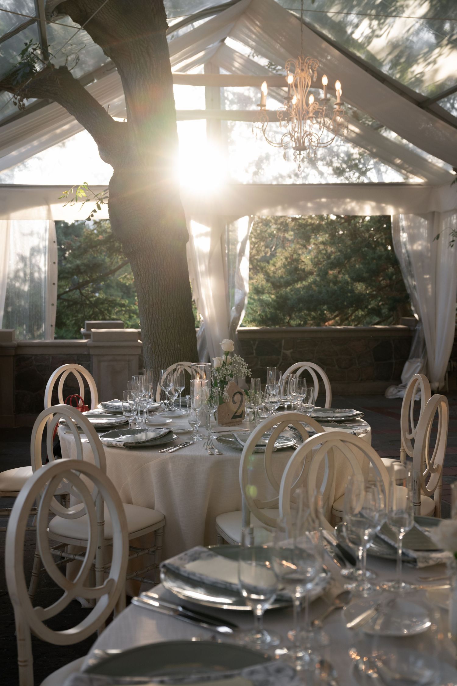 Elegant white wedding reception tables set with crystal glassware under a sunlit outdoor pavilion with flowing curtains.
