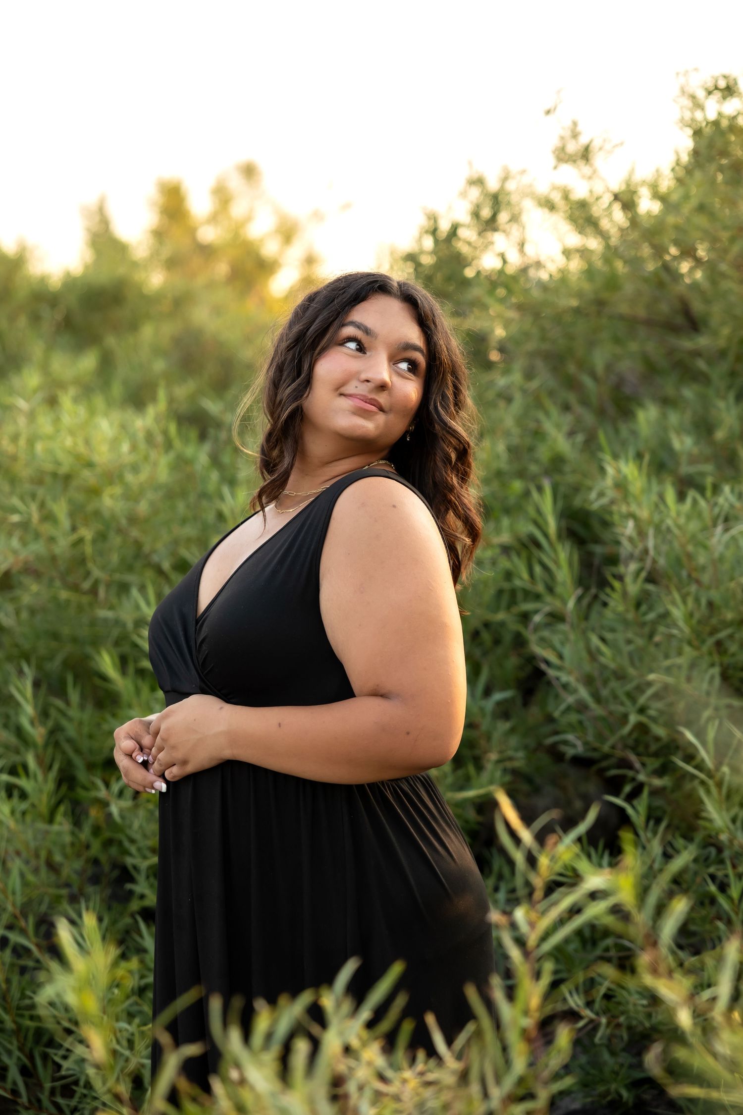 A woman in a black dress poses outdoors during golden hour in a field of tall grass and greenery.