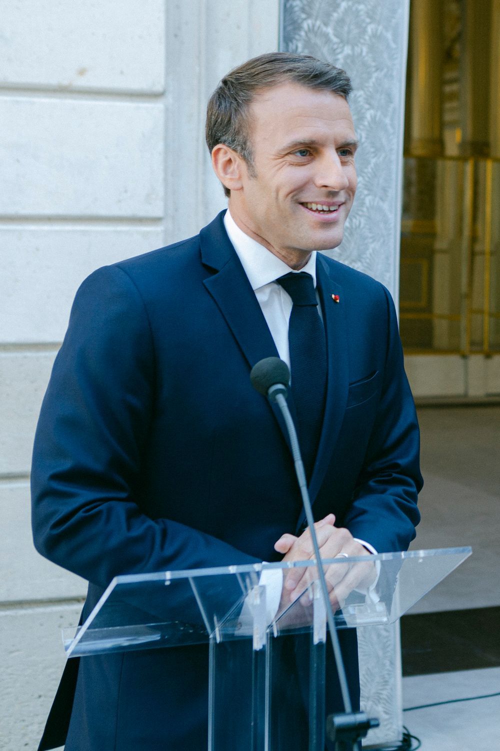 A person in a dark suit smiles while speaking at a podium with microphones during an official event.