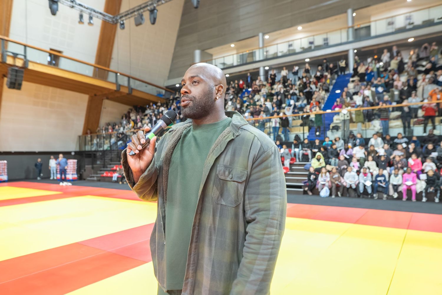 A person speaks into a microphone at a martial arts competition venue with spectators in stadium seating.