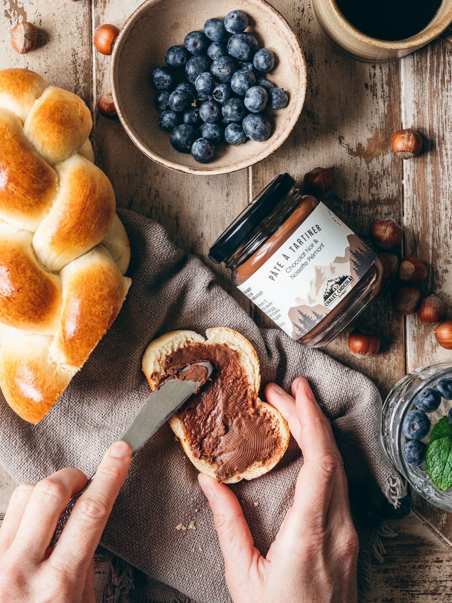Hands spreading chocolate spread on fresh bread alongside blueberries, coffee and hazelnuts on rustic wooden table.