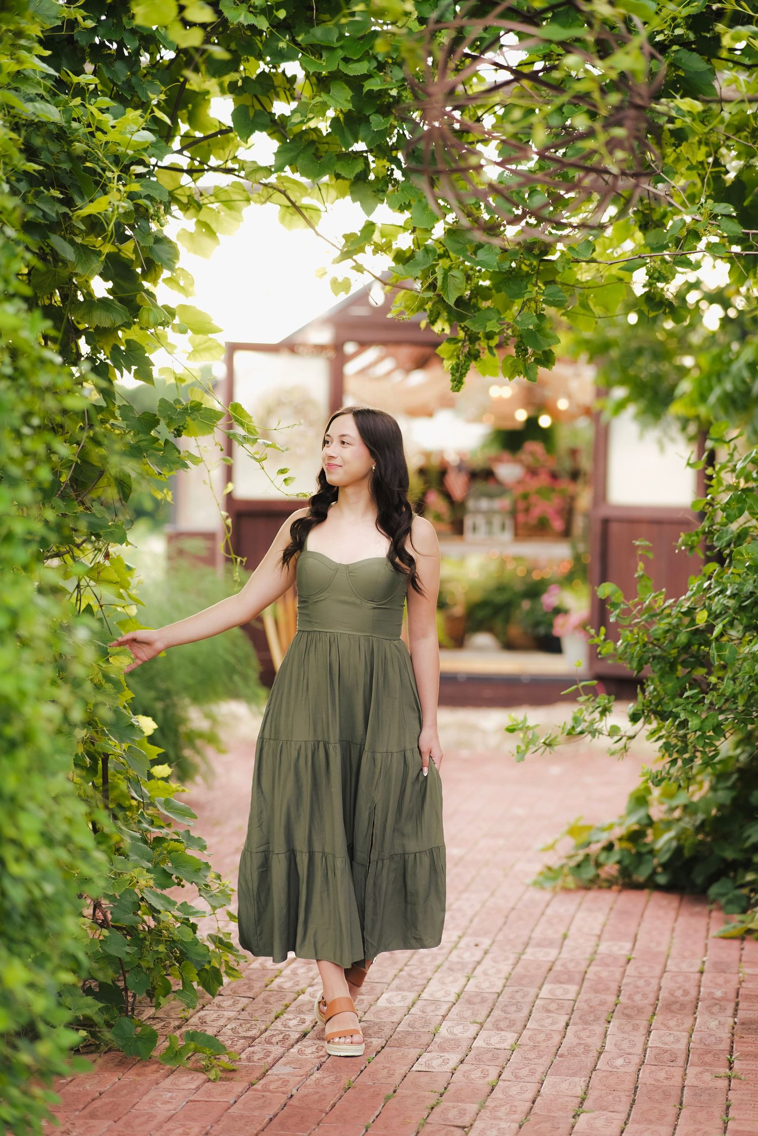 Young woman in an olive green maxi dress walks under a lush green vine-covered archway on a brick path.