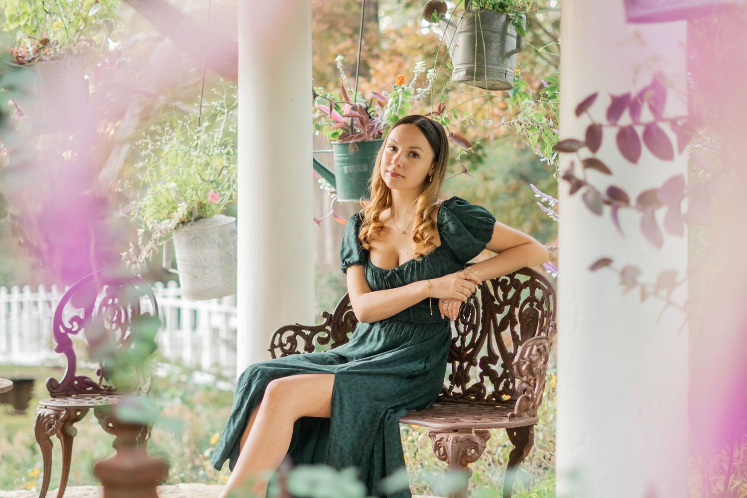 Young woman in dark green dress sits on ornate bench in a flower-filled garden with soft bokeh background.