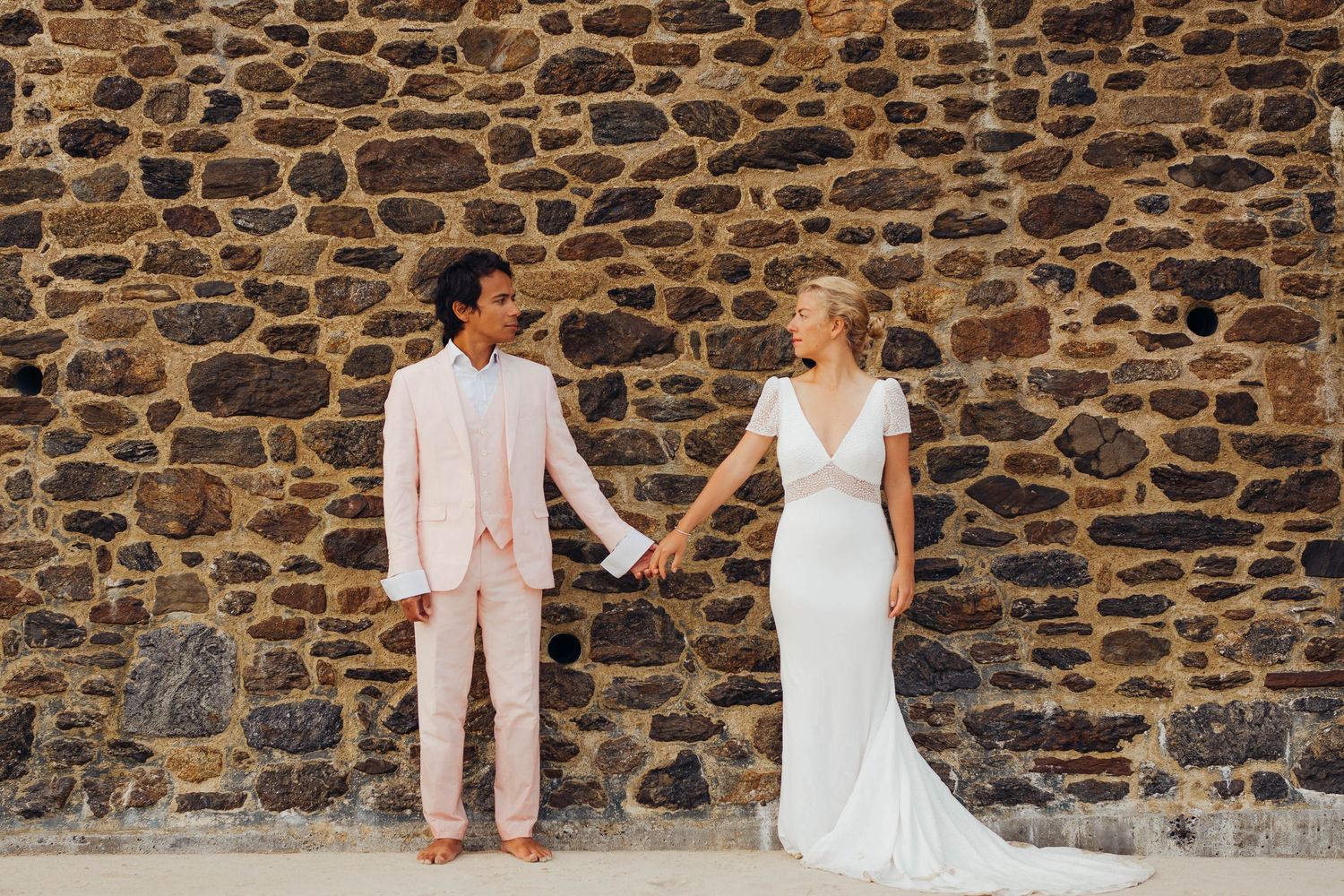 Bride in white gown and groom in pink suit hold hands before rustic stone wall outdoors.