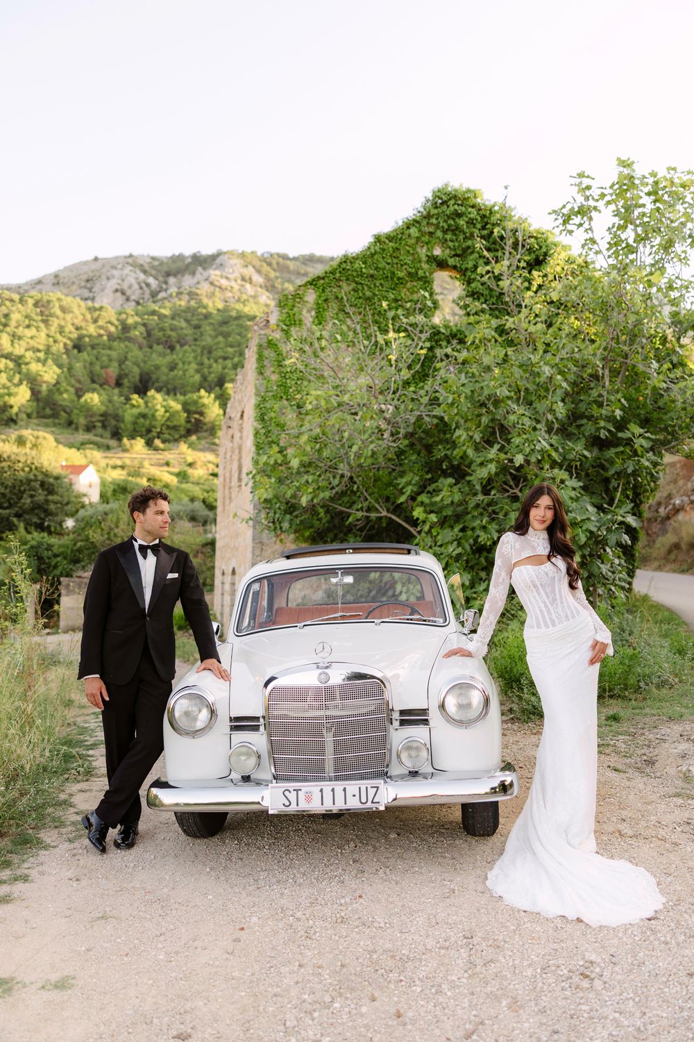 A classic Mercedes-Benz car parked in an orchard setting with people in formal attire during a wedding photoshoot, Hvar , Croatia