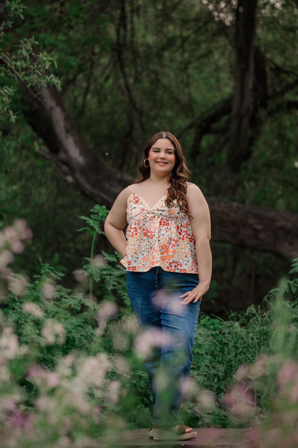 A floral top and jeans outfit photographed in a lush forest setting with pink wildflowers in the foreground.