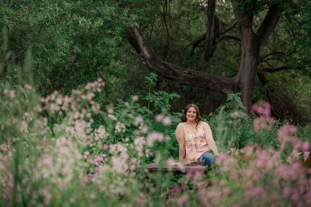 A woman in a floral dress poses in a lush green garden setting with pink wildflowers blooming around her.