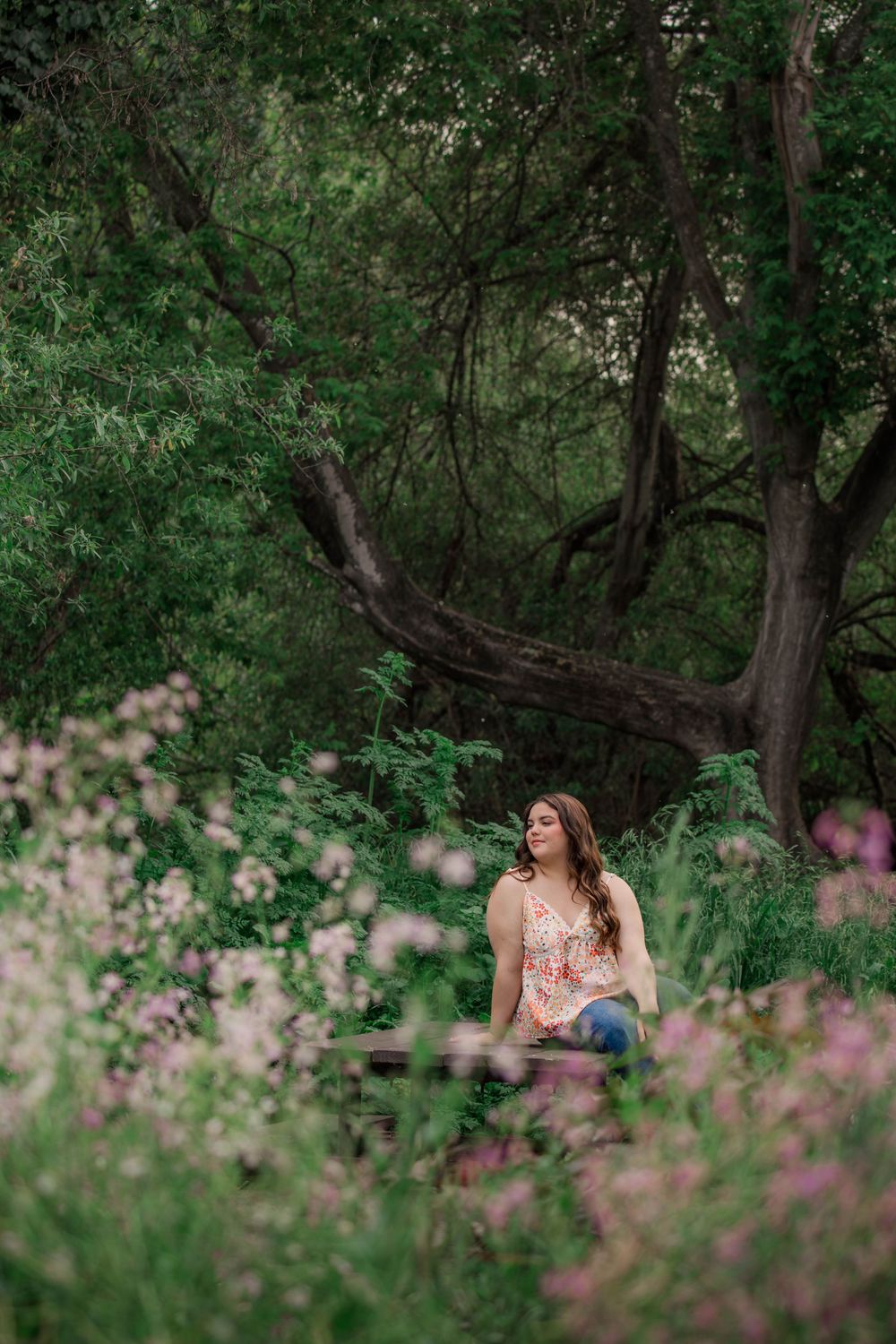 A person sits peacefully in a garden setting surrounded by pink wildflowers and lush green trees on a summer day.