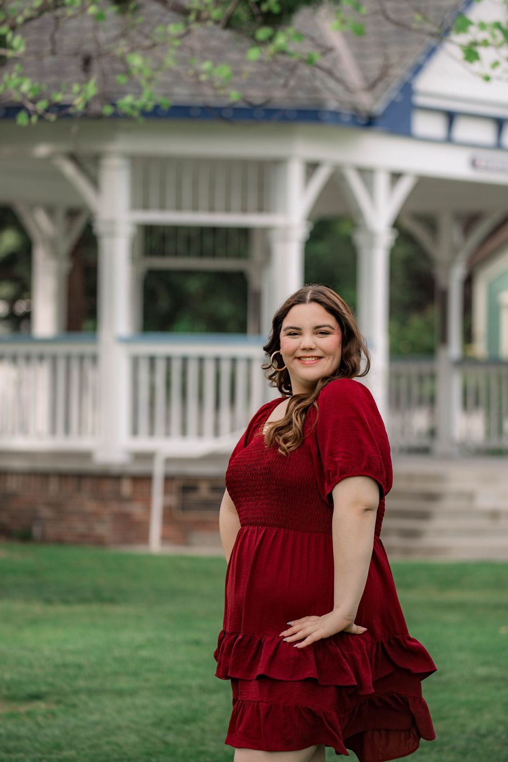 A person in a burgundy ruffled dress poses outdoors near a white gazebo and green lawn.