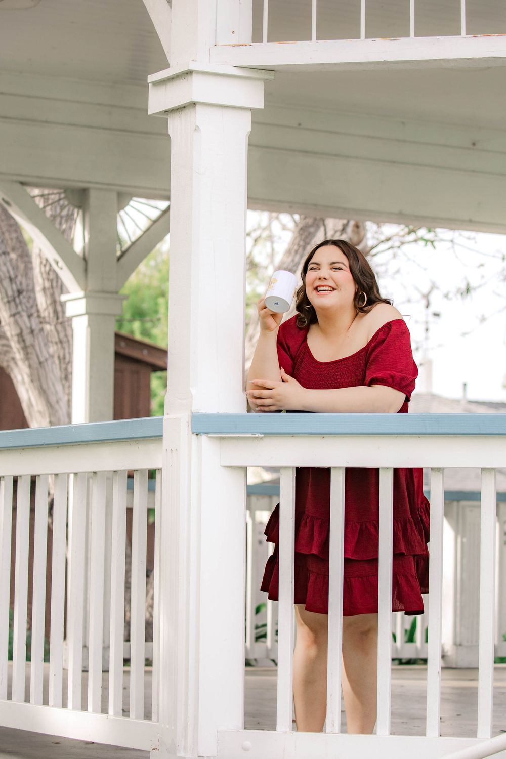 Woman in red dress leans on white porch railing while laughing and enjoying a sunny day outdoors.