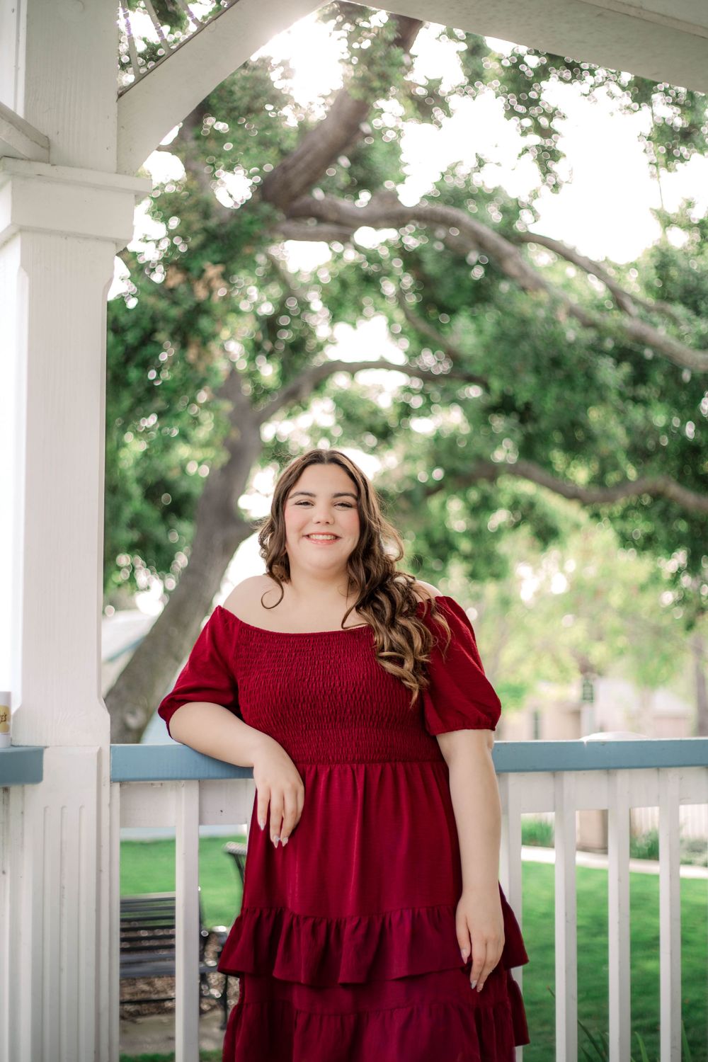 A person in a burgundy dress smiles while standing in a white gazebo with greenery in the background.