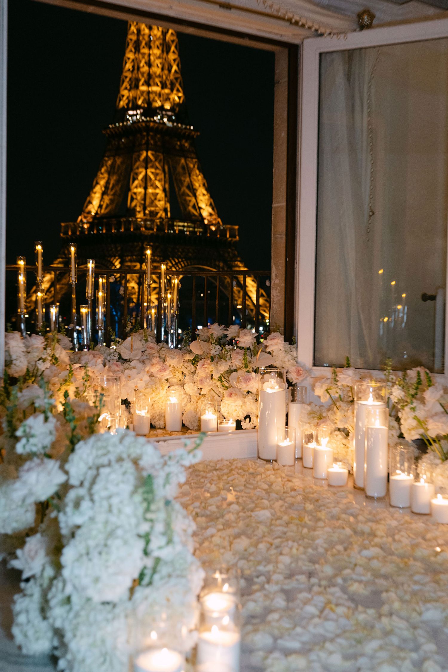 White floral arrangements and glowing candles create a romantic ambiance with the illuminated Eiffel Tower visible through windows.