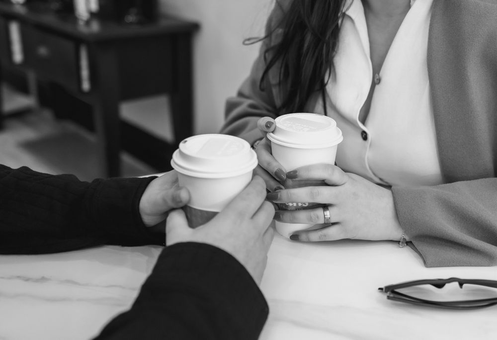 Business people sitting at tables with coffee cups and laptops in modern office setting.