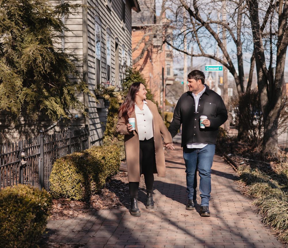 A romantic couple walks together on a path surrounded by autumn trees and stone architecture.
