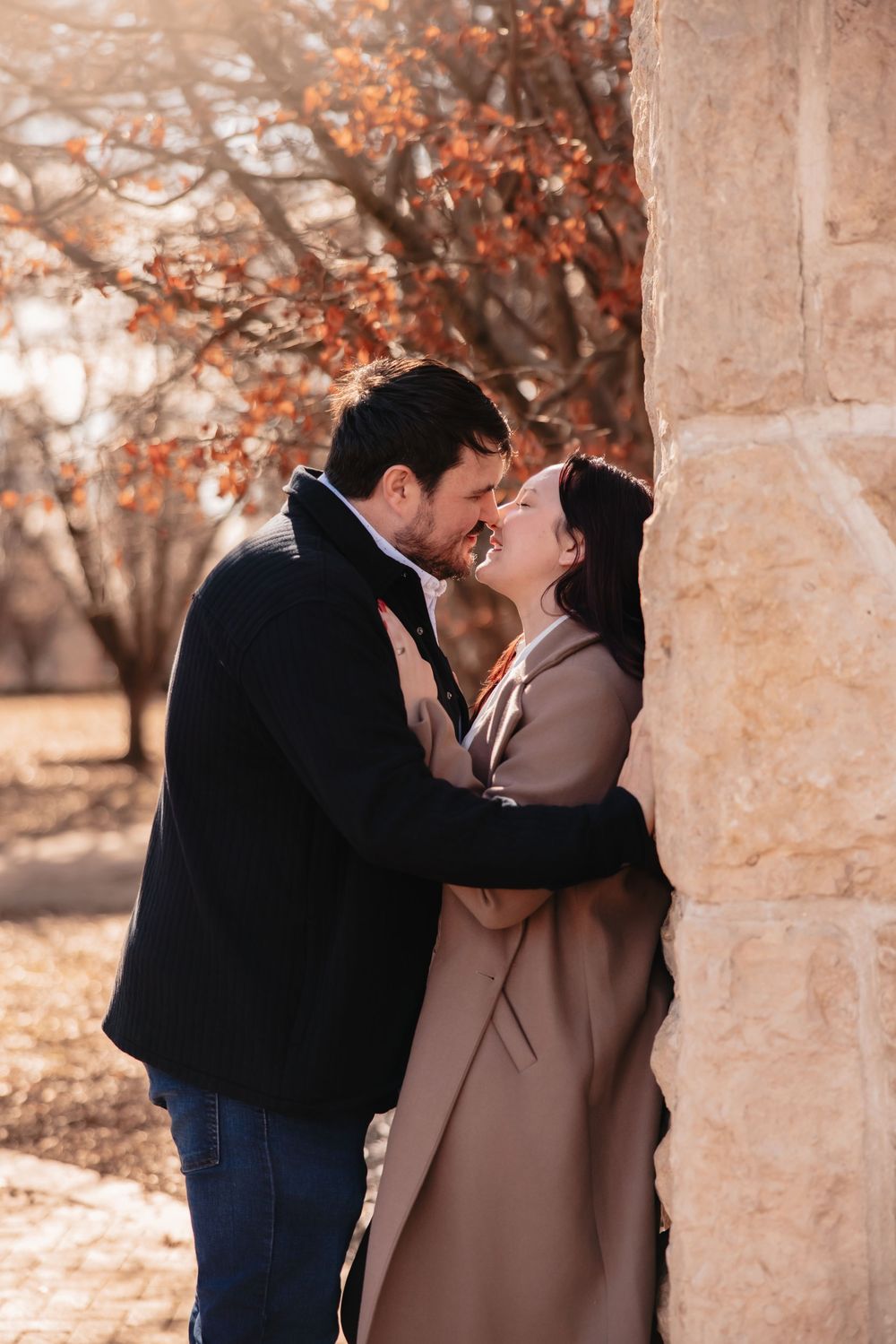 A couple shares an intimate moment against a stone pillar backdrop on a fall day.