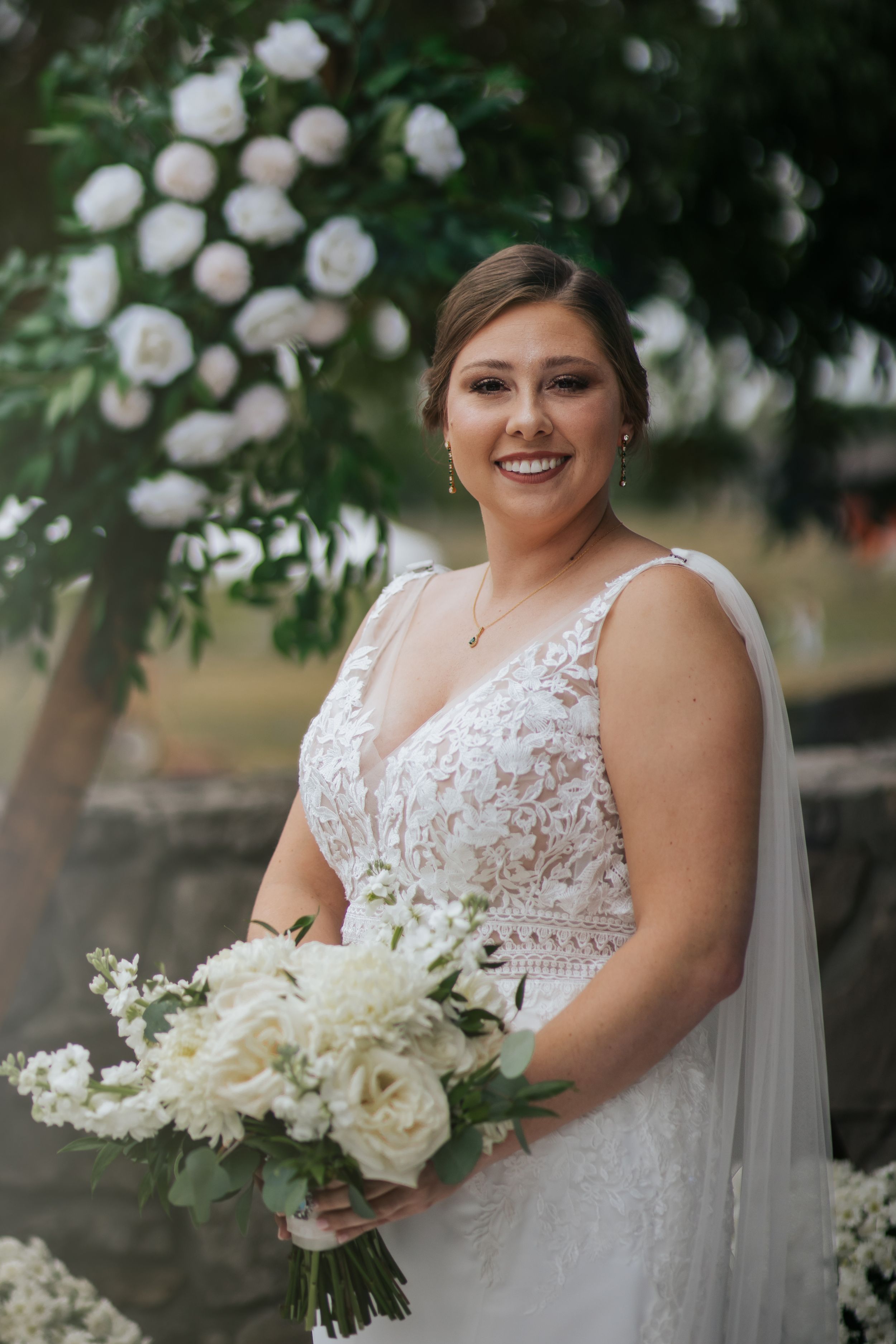 Bride in lace wedding dress holding elegant white floral bouquet poses under blooming trees in outdoor setting. Conner Prairie, Fishers Indi