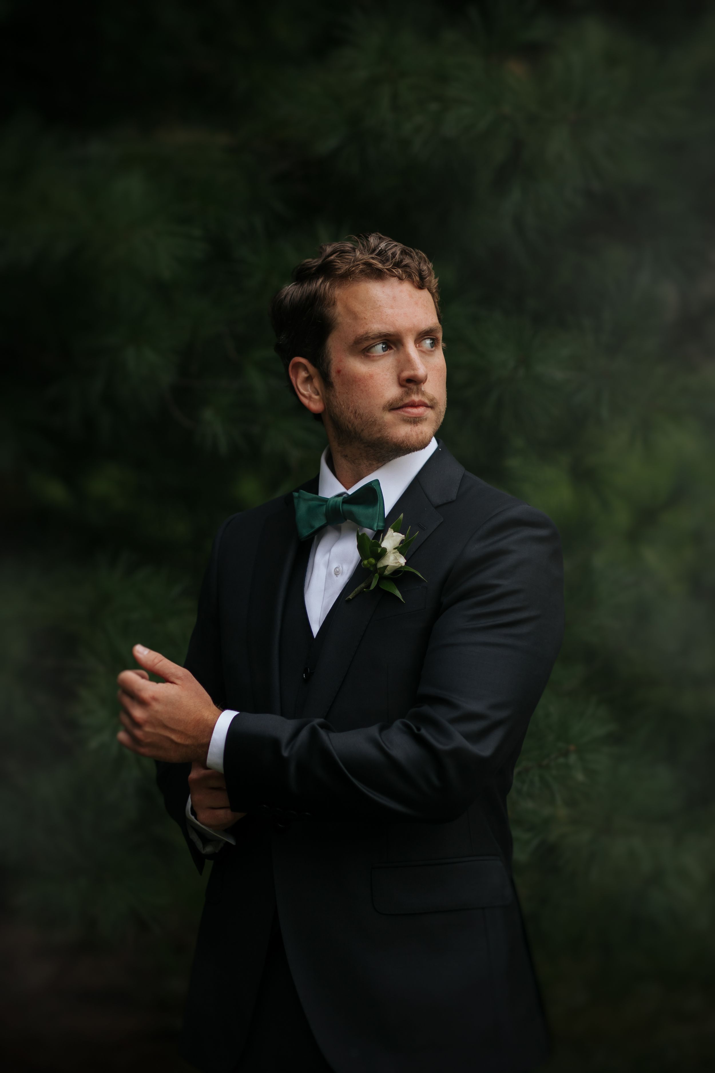 A man in a black tuxedo adjusts his tie while standing against a dark background in a formal portrait series. Conner Prairie, Fishers Indian