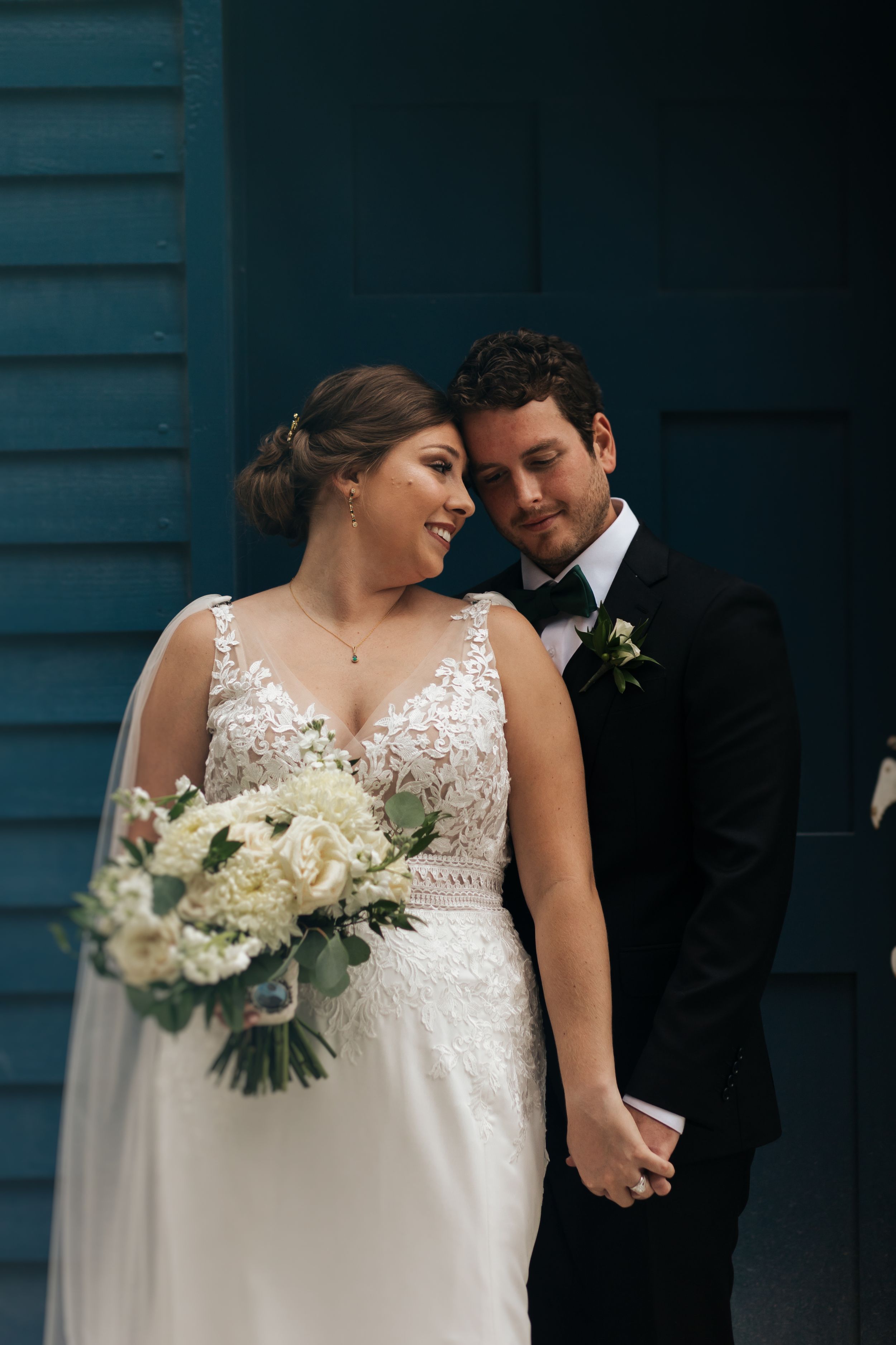 Intimate wedding portrait against blue wooden wall with white floral bouquet.