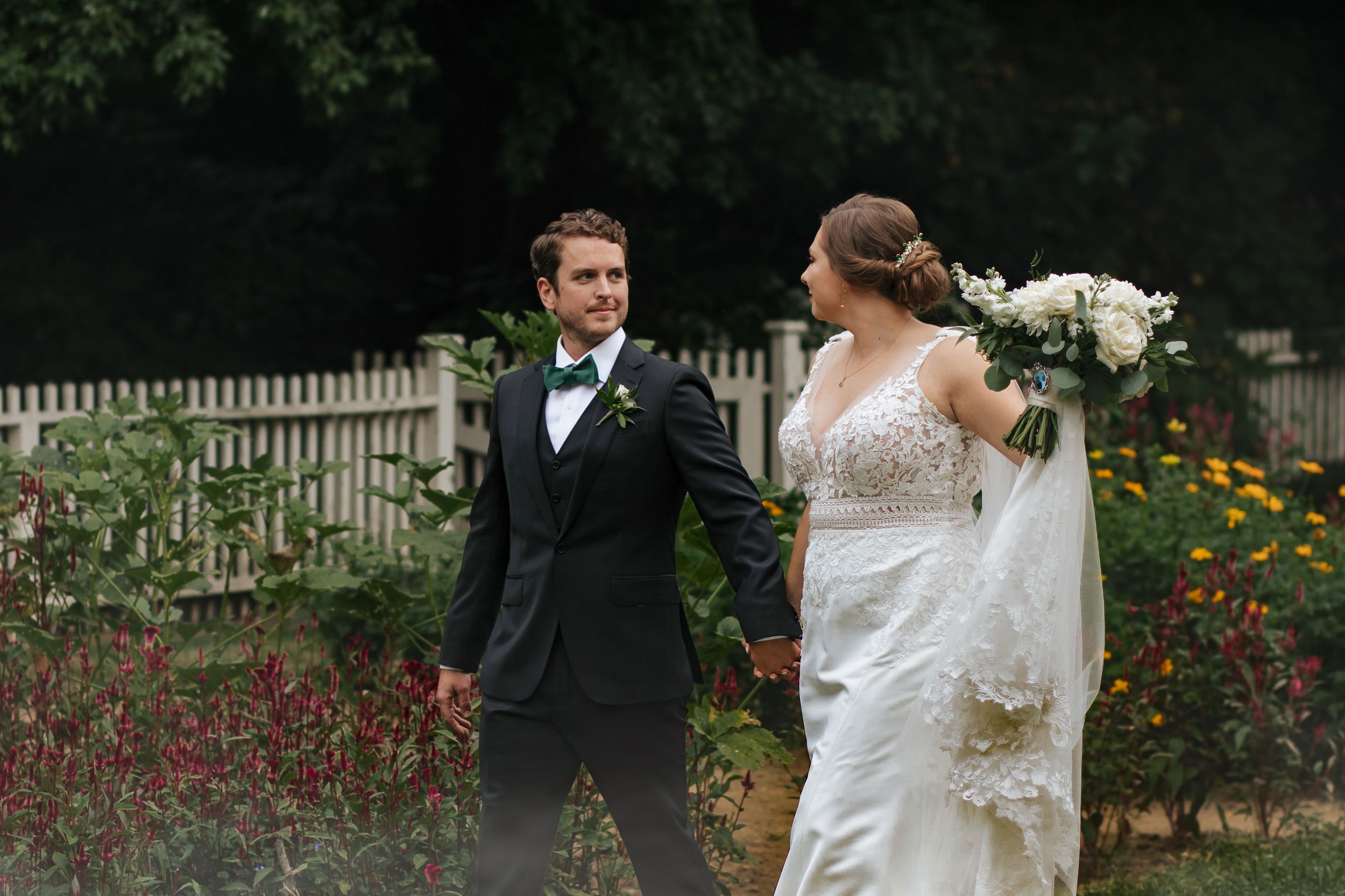 A wedding couple poses in a garden with white picket fence and orange flowers in the background.