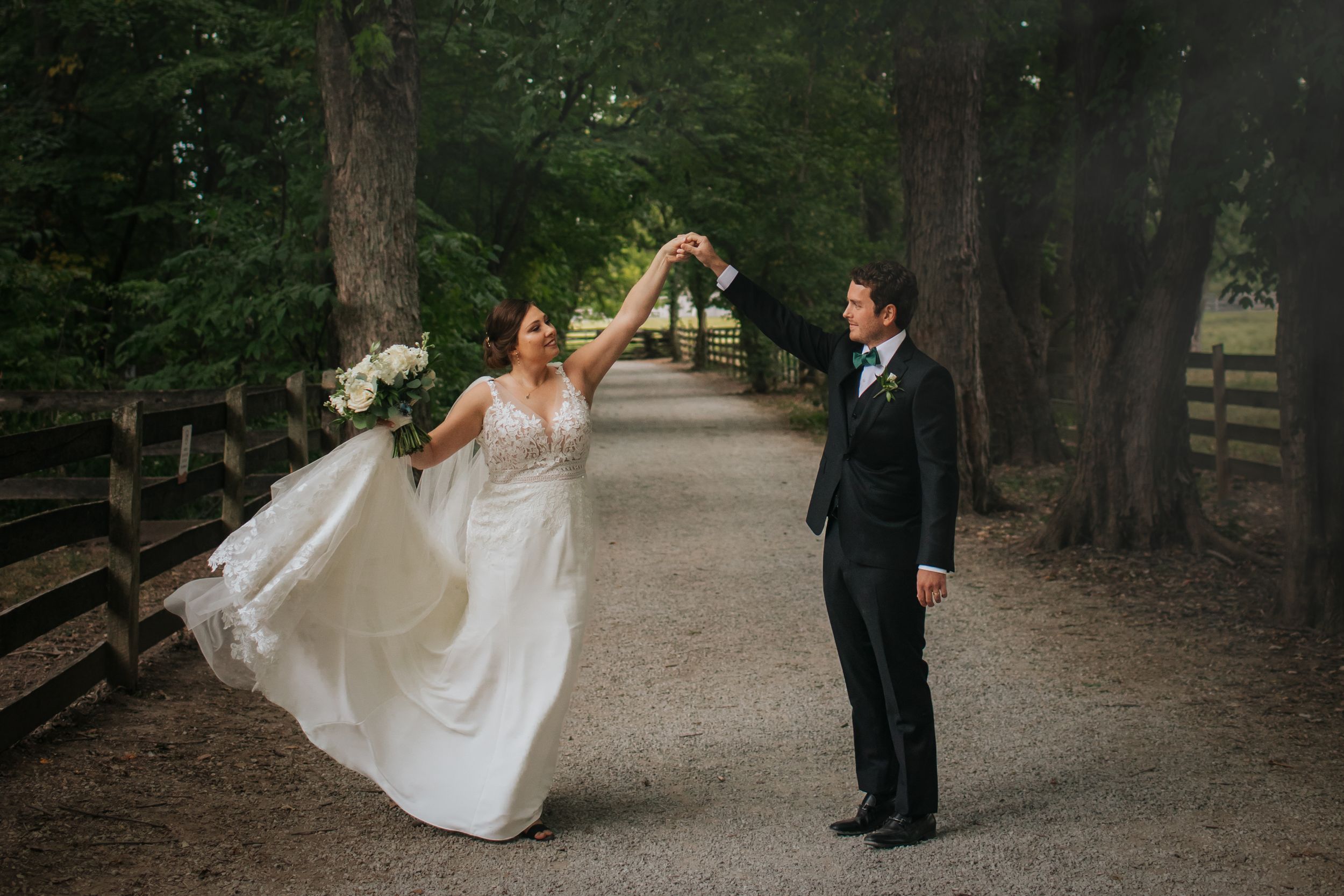 A playful wedding moment captured on a tree-lined path with white dress flowing.