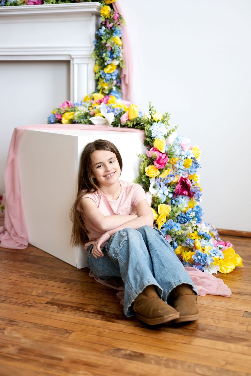 Smiling girl in pink shirt and jeans sitting beside a white cube with colorful spring floral decor.