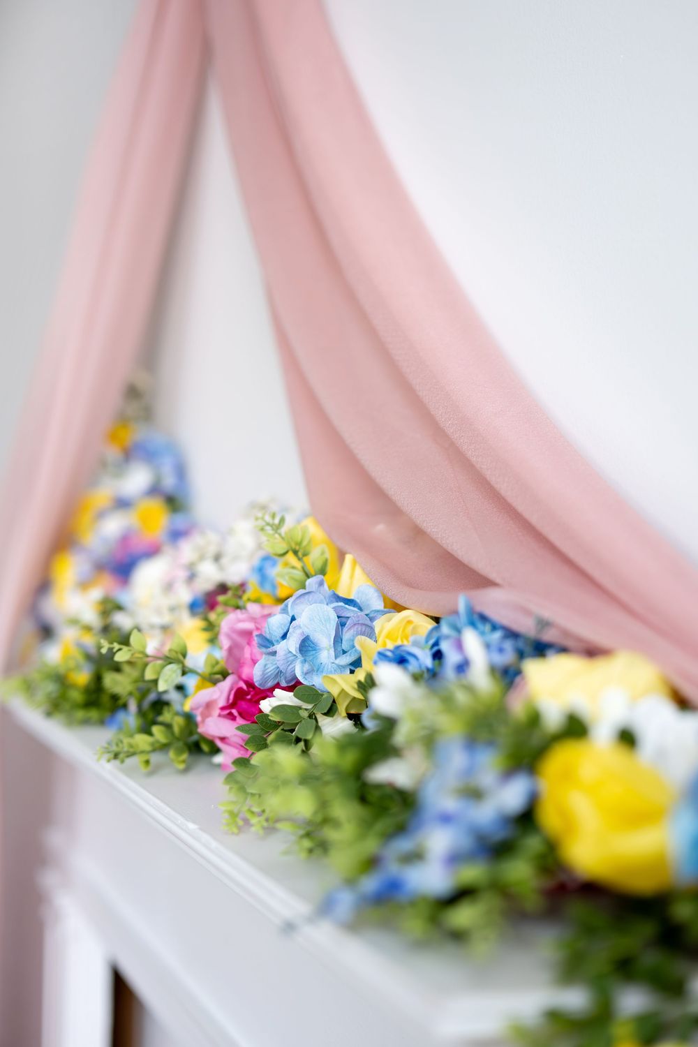 Colorful spring flower garland with blue, pink, and yellow blooms on a white mantel with draped pink fabric.