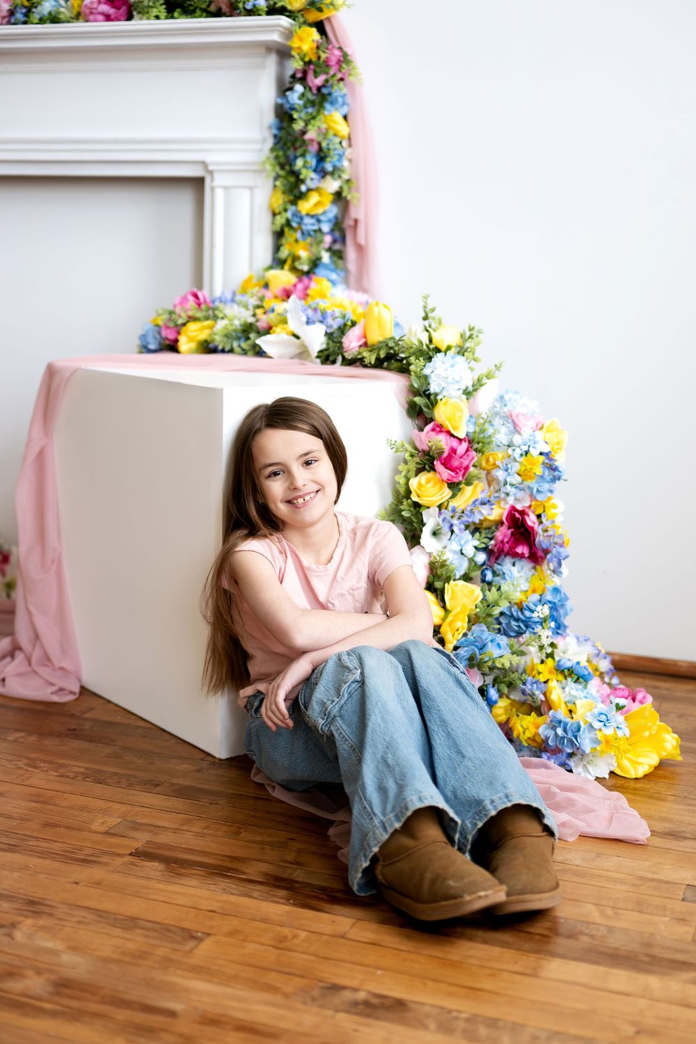 Cheerful girl in jeans and pink top posing beside white cube with colorful floral spring backdrop.