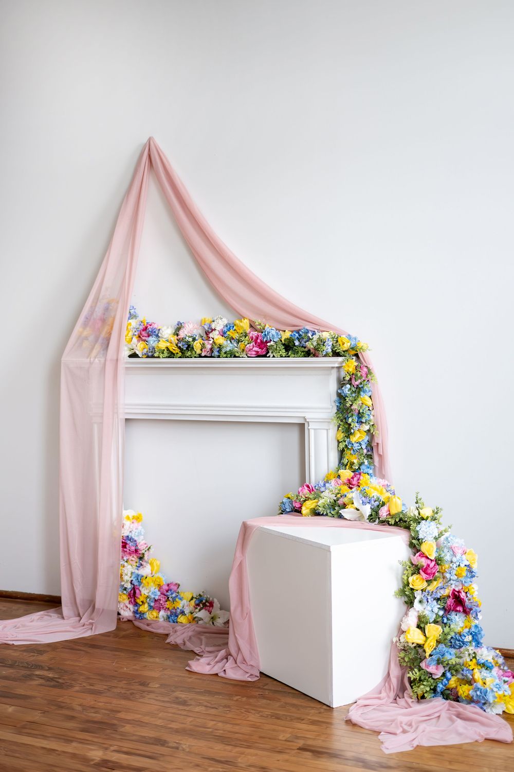 White decorative fireplace mantel adorned with colorful flower garlands and flowing pink fabric draping to the floor.