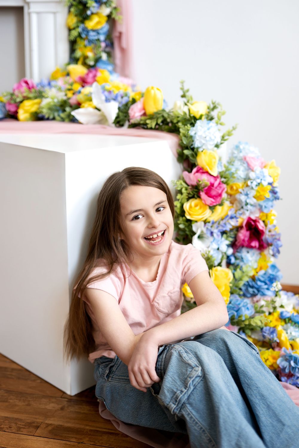 Laughing girl in pink shirt and jeans seated on floor with colorful spring floral mantel backdrop.