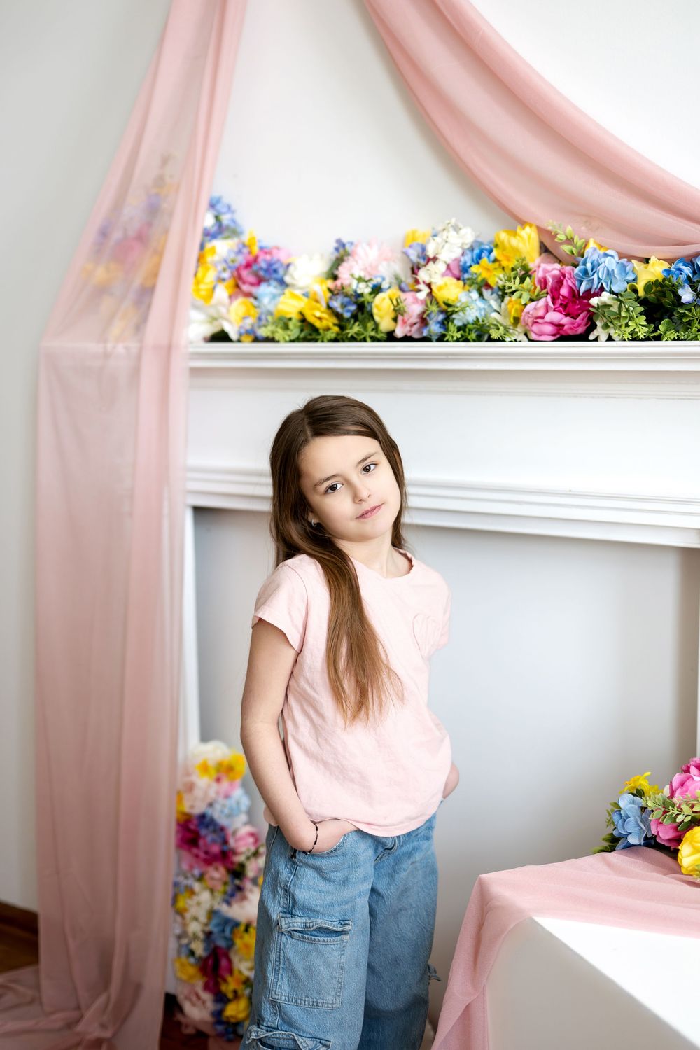 Young girl in pink tee and jeans standing before white fireplace adorned with colorful spring flowers.