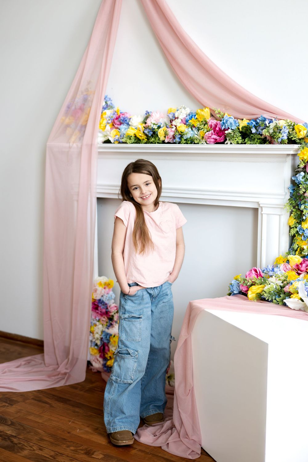 Smiling girl in pink shirt posing beside flower-decorated white fireplace with pink sheer fabric canopy.