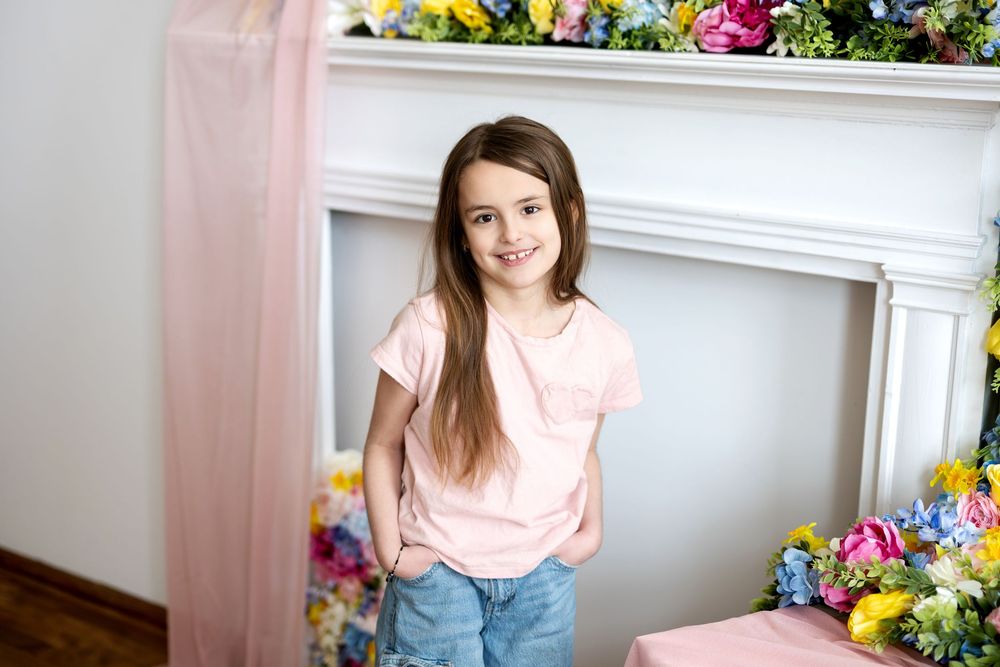 A young girl in a pink t-shirt and jeans smiles in front of a colorful flower-adorned fireplace mantel.