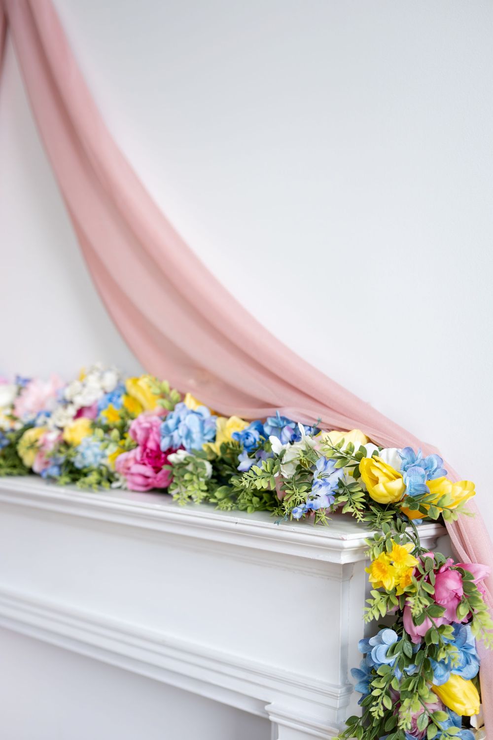 White fireplace mantel decorated with colorful garland of pink, yellow, blue flowers and draped pink chiffon fabric.