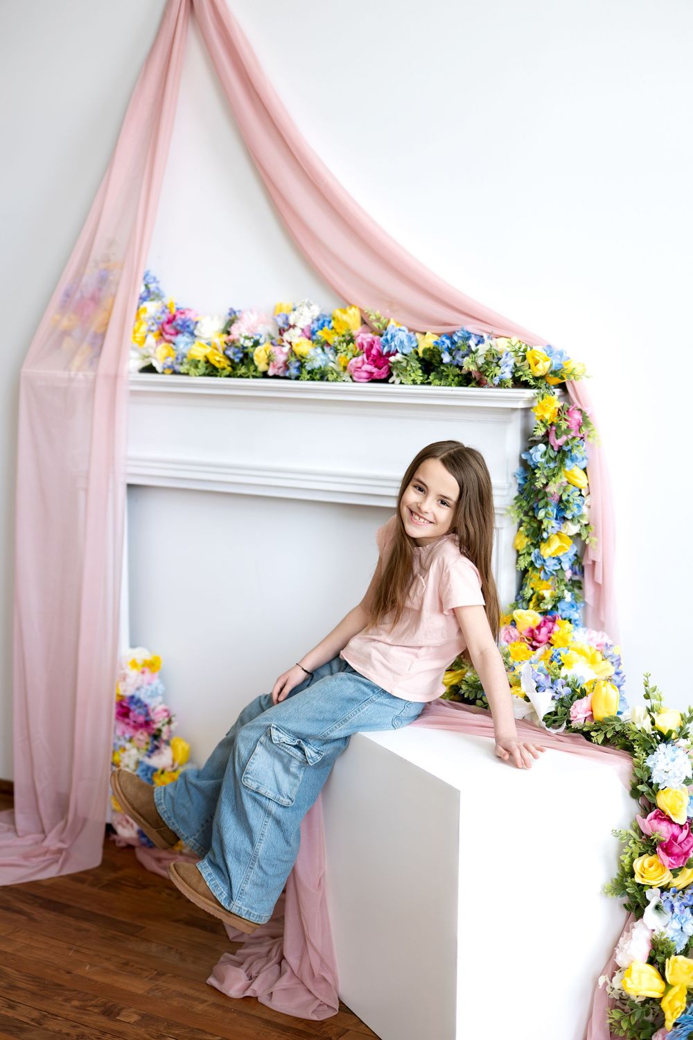 A cheerful girl in jeans and pink tee sits on a white cube beside a flower-decorated fireplace with pink draping.