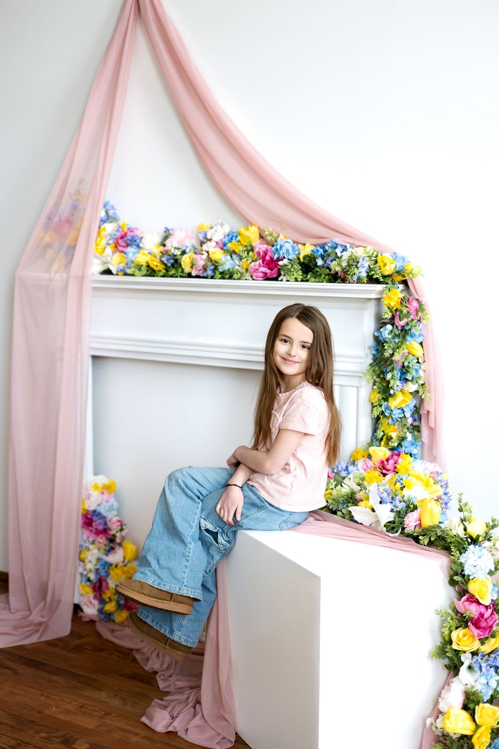 A young girl in a pink shirt and blue jeans sits smiling on a white cube by a flower-decorated fireplace.