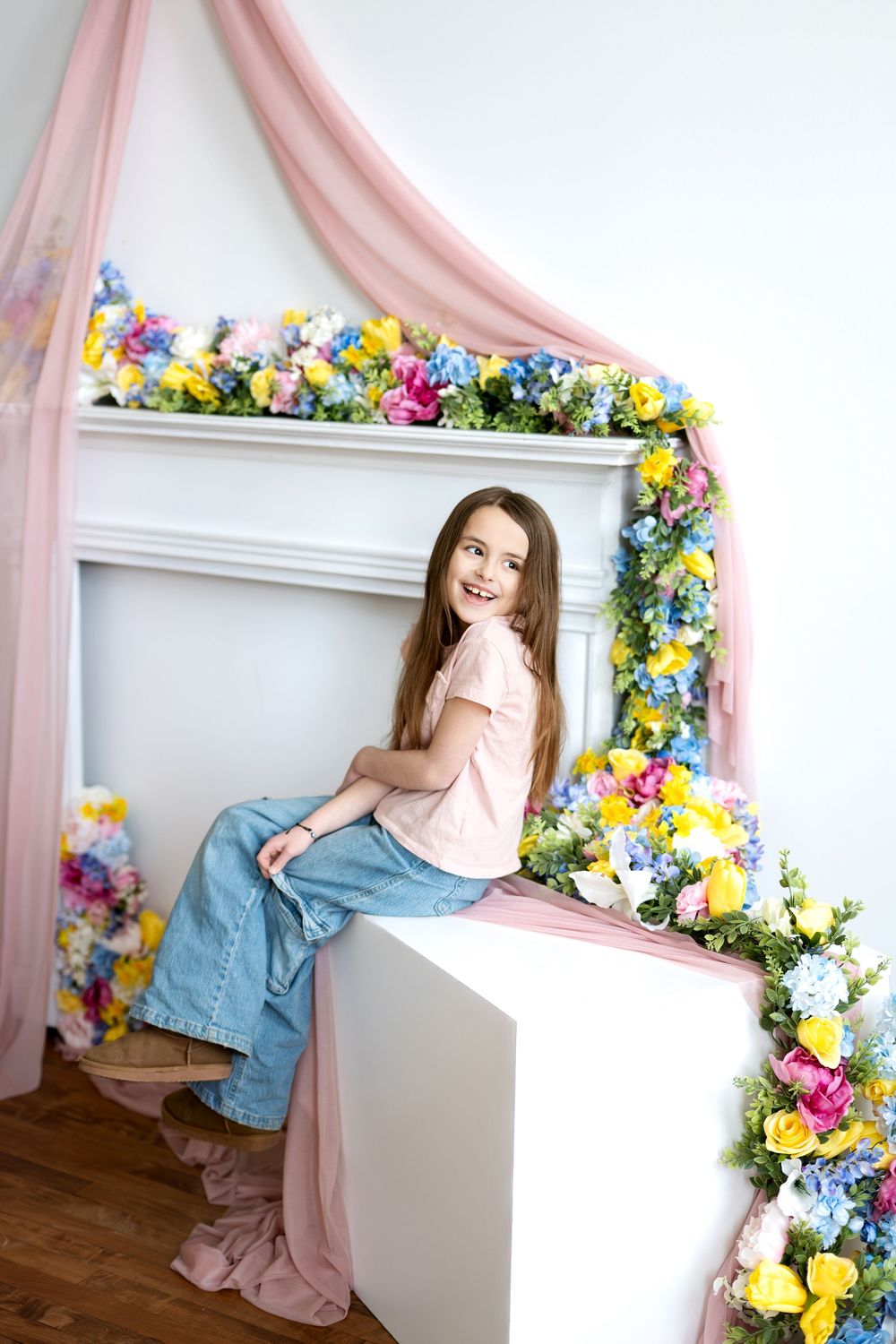 A laughing girl in jeans and a pink tee sits on a white cube beside a vibrant flower-decorated fireplace.