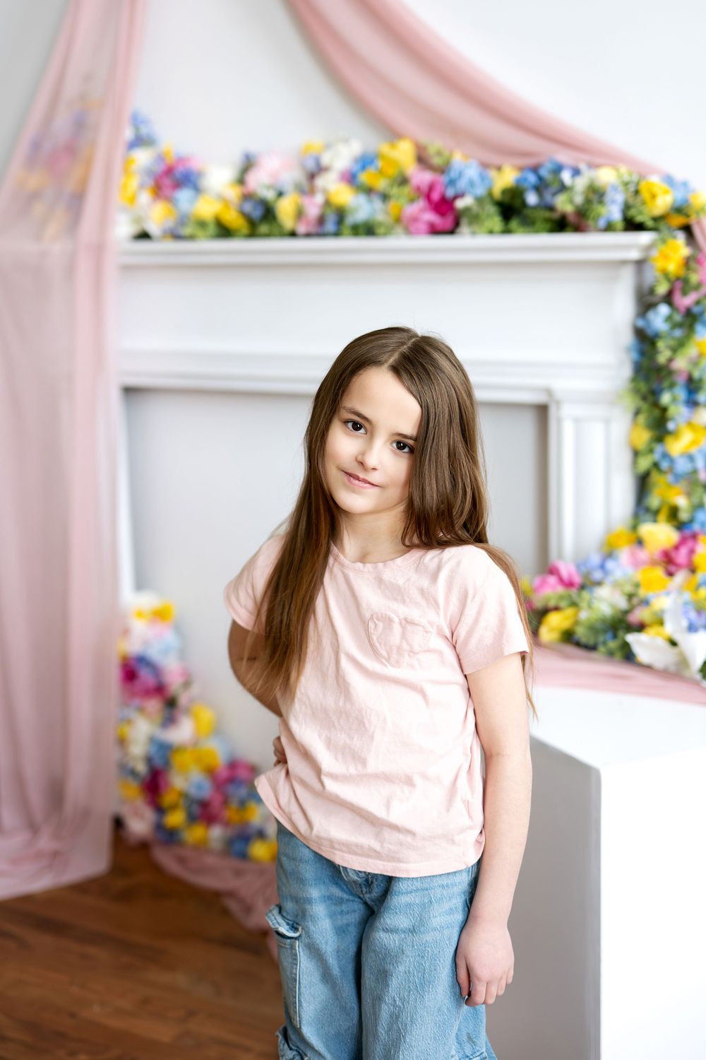 A young girl with long brown hair poses in a pink t-shirt and jeans before a floral-decorated white fireplace.
