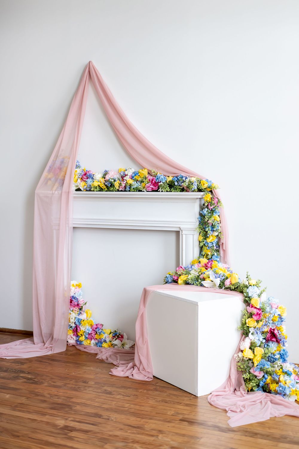 White decorative fireplace adorned with colorful spring flowers and flowing pink tulle fabric draping.